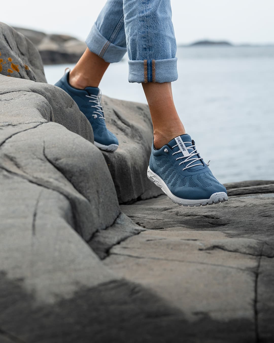 Person in rolled-up jeans walking on cliffs by the sea, wearing Icebug Eli RB9X blue sneakers.