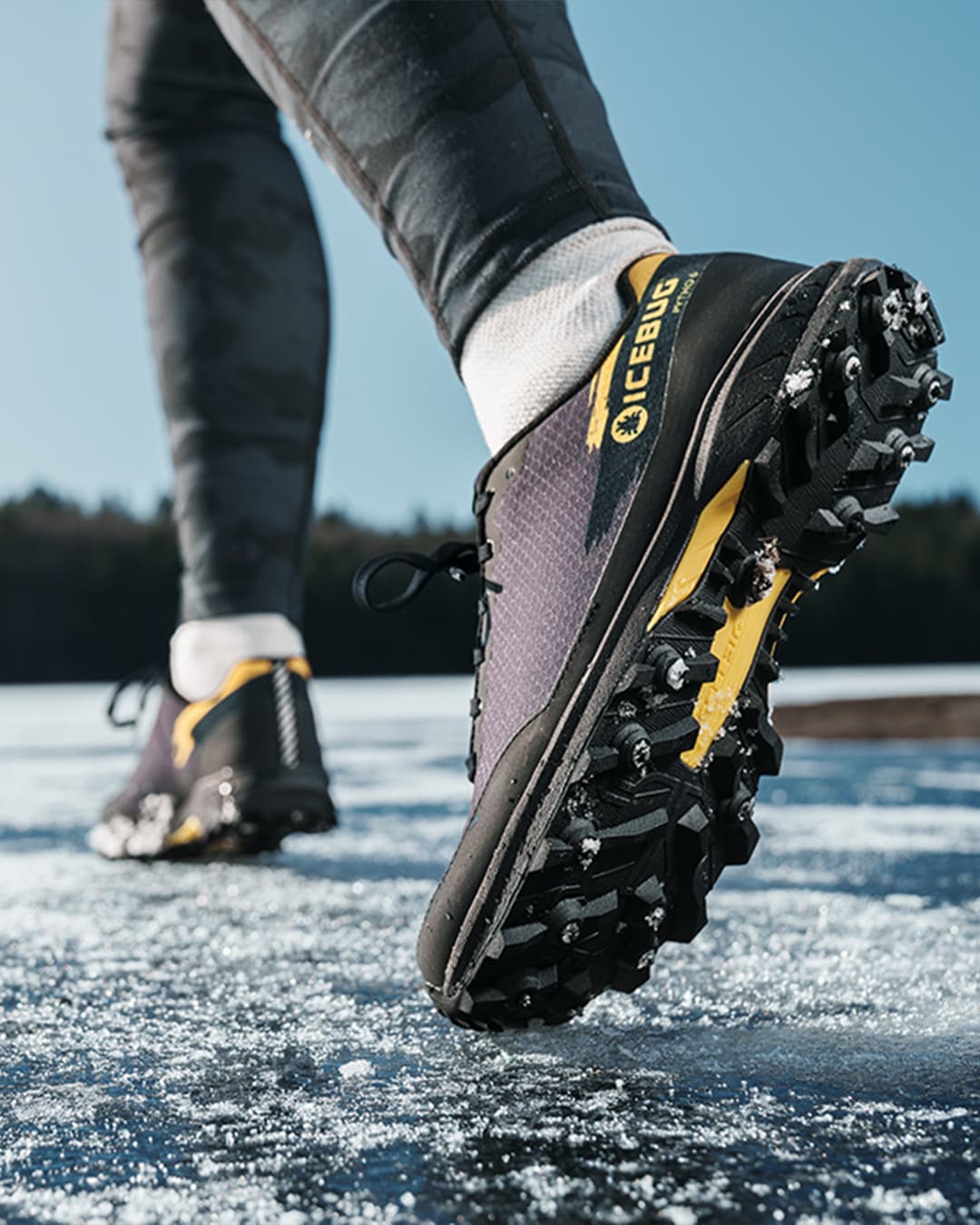 Close-up of a person running on icy ground wearing studded Icebug trail running shoes, with a clear sky and forest in the background.