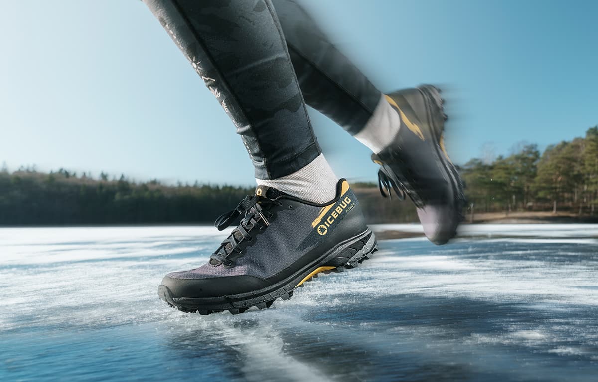 Close-up of a person running on ice in black and yellow studded trail running shoes from Icebug, with blurred motion. Forest and clear sky in the background.
