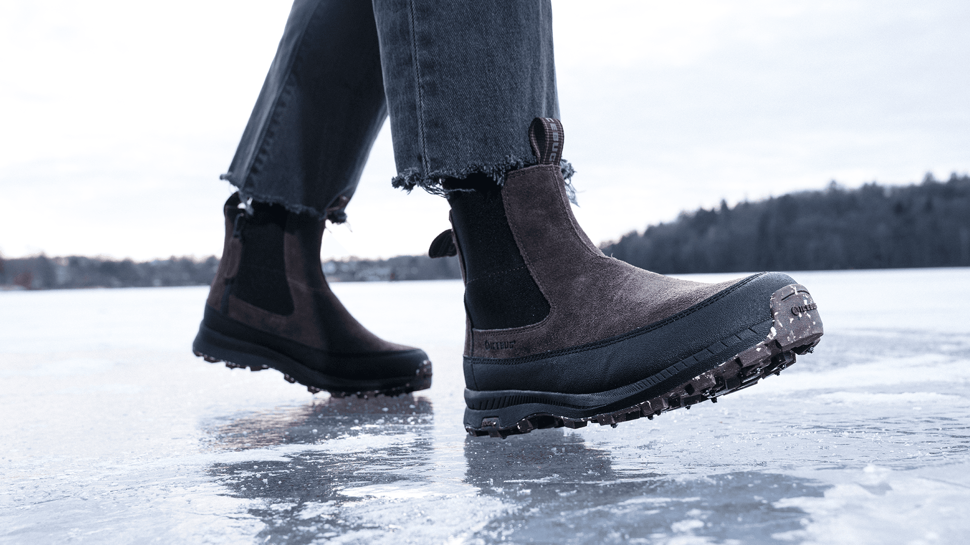 Person wearing brown studded winter boots with black soles walking on ice, wearing frayed black jeans; forest visible in the background.