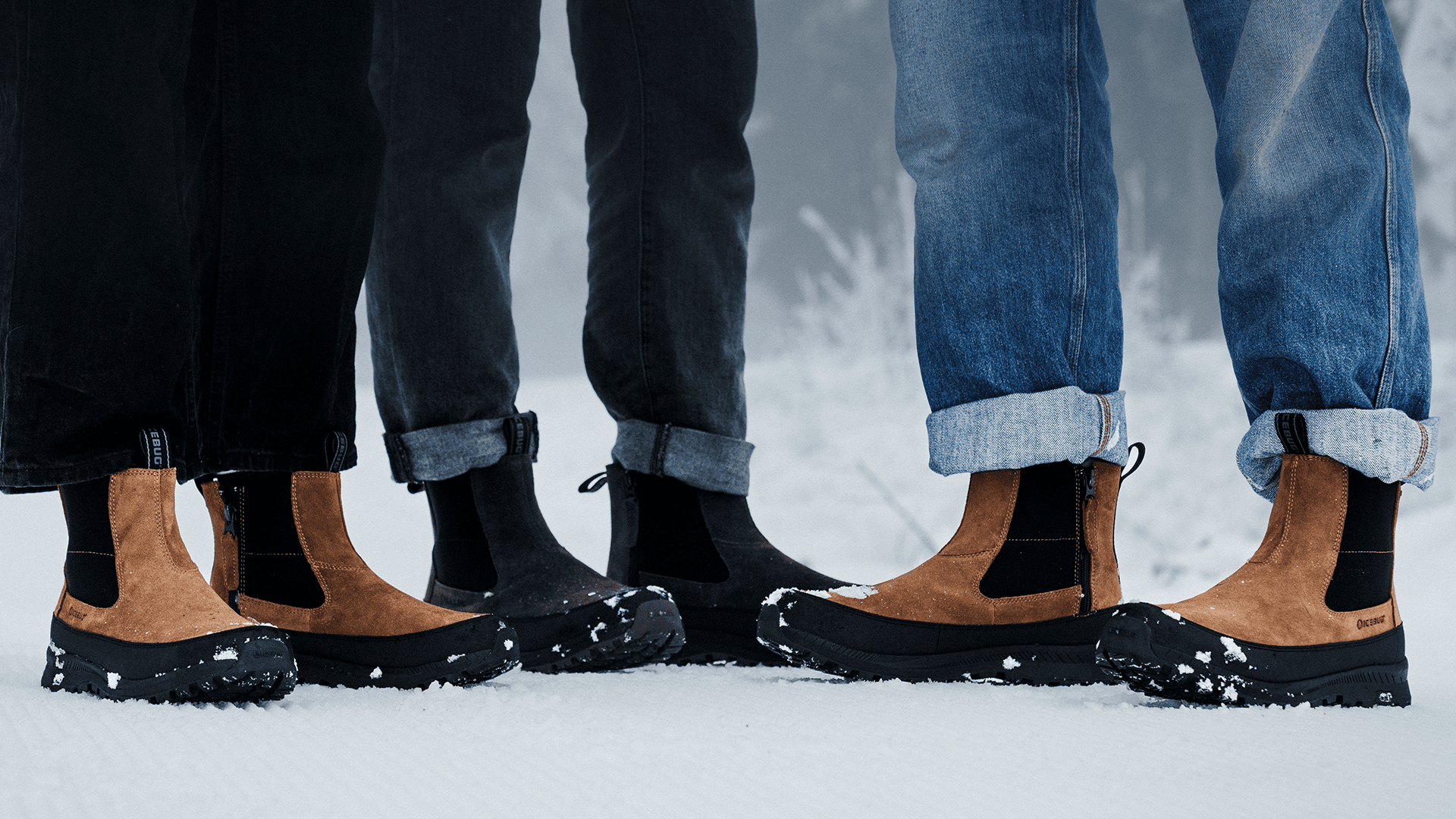 Three people wearing brown and black Icebug winter boots stand on snow, with jeans rolled up at the cuffs.