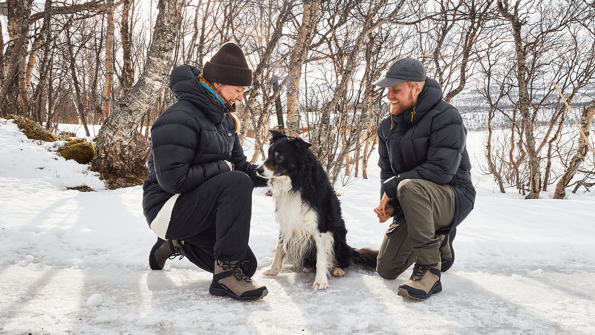 A man and woman in winter clothing kneel beside a black and white dog on a snowy path, surrounded by bare trees.