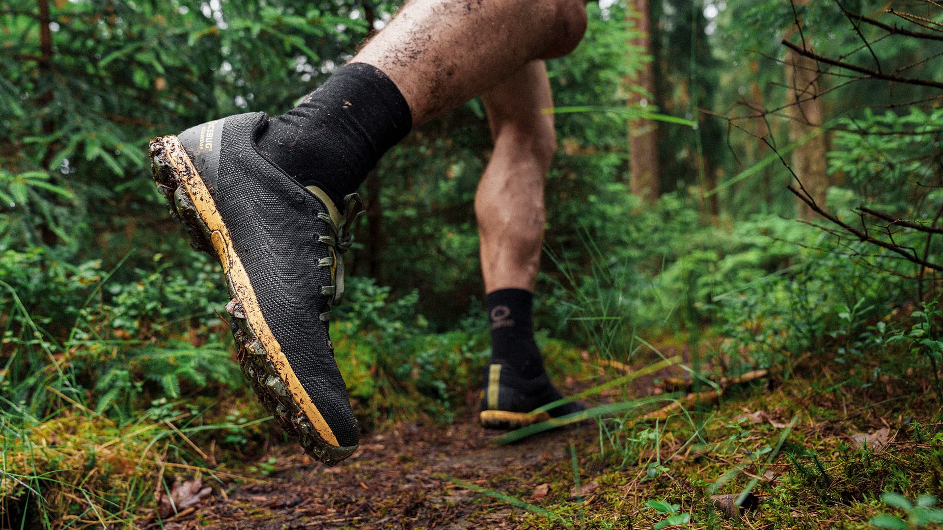 Close-up of a person trail running in a forest, wearing black shoes with muddy soles. Lush greenery surrounds the path.