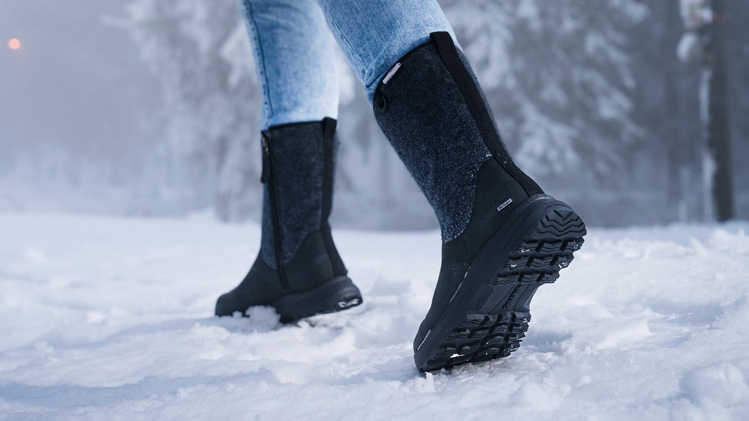 Person wearing tall black winter boots and light blue jeans walking on snow, with a foggy forest background.
