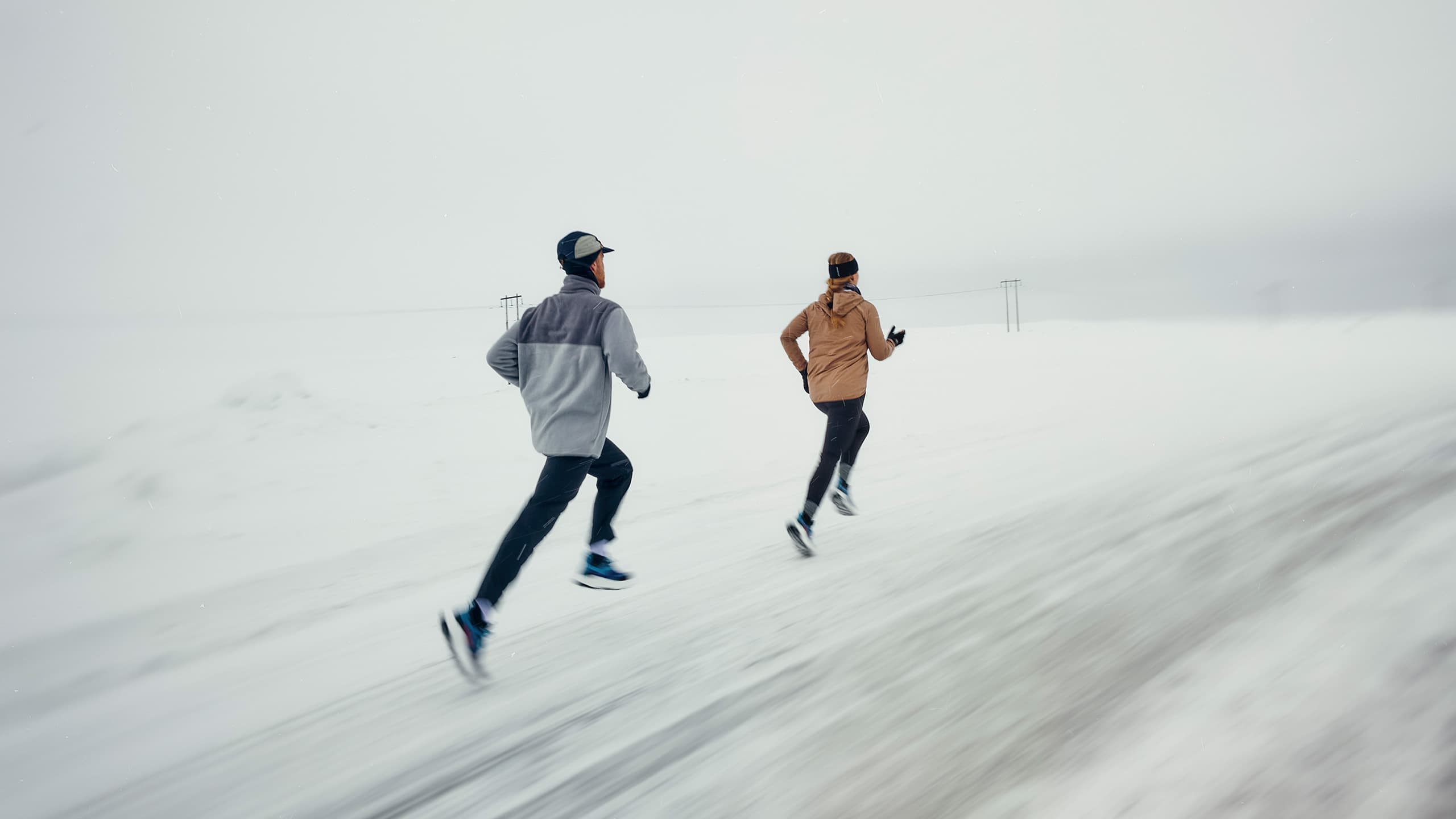 Two people running on a snow-covered road, wearing winter clothing. The sky is overcast, and the landscape is a vast snowy expanse.