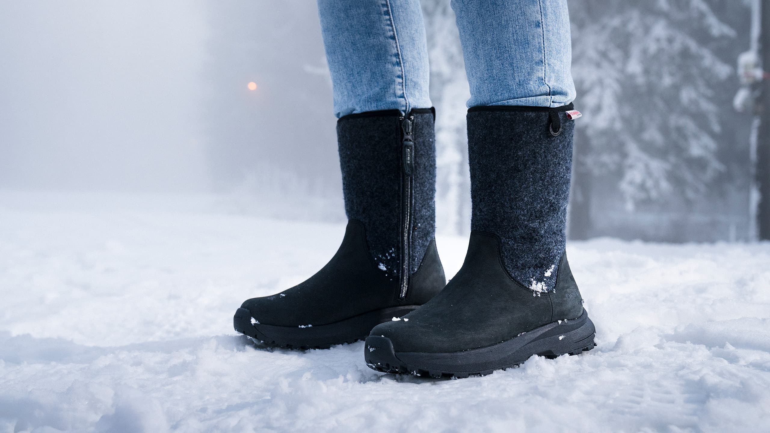 Person in blue jeans wearing black winter boots stands in snow, with a foggy, snowy background.